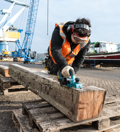 Man met veiligheidsbril schuurt een grote biels. Op de achtergrond een scheepswerf.